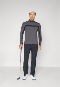 Gray zip-up jacket with black accents, paired with black pants and white golf shoes. The model holds a golf club and wears a white cap.