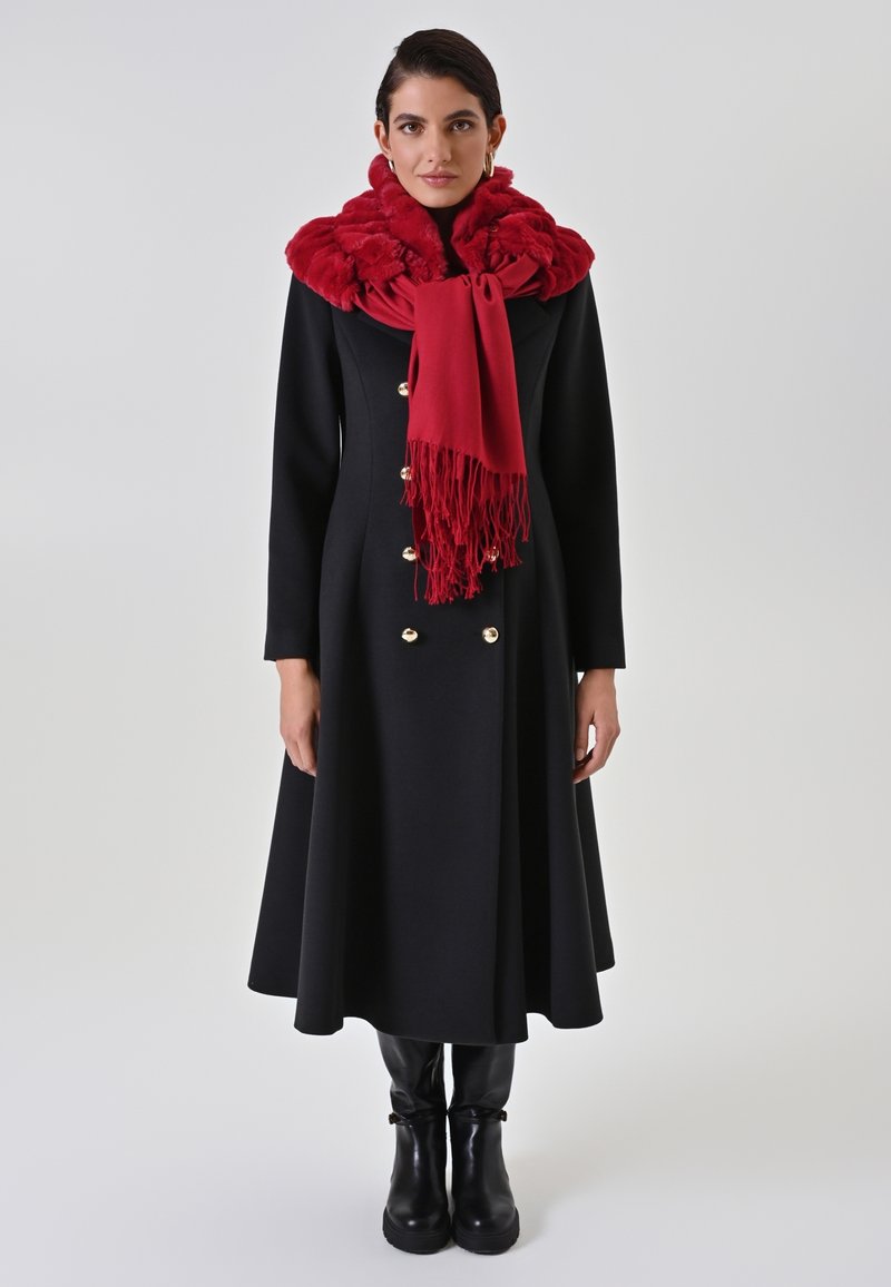 Cappotto nero con bottoni dorati e design doppiopetto, abbinato a una sciarpa rossa strutturata con frange. Texture liscia e silhouette aderente.
