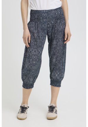 Woman wearing navy blue paisley-patterned cropped pants with smocked waist and cuffs, white top, and beige sneakers with black stripes.