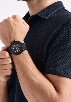 Moški nosi črno teksturirano polo majico in prikazuje črn kronograf z rdečimi detajli na zapestju proti beli ozadju.