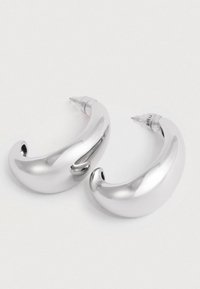 Para masywnych, wypolerowanych srebrnych kolczyków w kształcie kół z zapięciem na sztyftach, leżących na gładkiej białej powierzchni.