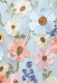 Viacfarebné vyšívané kvety, vrátane modrých, ružových, bielych a oranžových okvetných lístkov s hnedými stredmi, na priesvitnej svetlomodrej látke so zelenými listami.