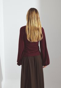 Bordo rebrasti dolg rokav z utesnjenim pasom in zvončastimi rokavi, v kombinaciji z rjavo krilom visoko v pasu, vidno s hrbta.