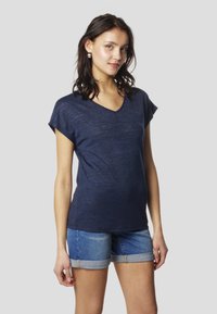Tmavomodré tričko s krátkym rukávom a výstrihom do V z ľahkej tkaniny, s jemnou textúrou a vyšívaným logom na prednej strane, v kombinácii s rifľovými šortkami.