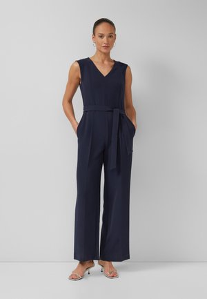 Femme portant une combinaison sans manches bleu marine avec col en V, taille ceinturée, jambes larges et talons argentés à bouts ouverts, debout contre un mur gris uni.