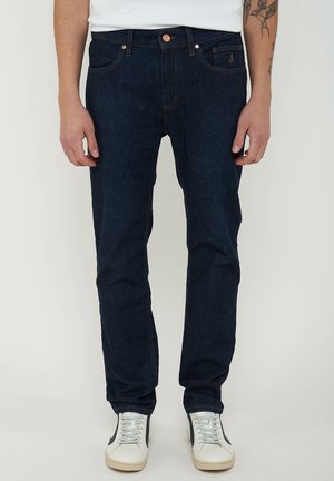 Man draagt donkerblauwe rechte jeans en witte sneakers, staande tegen een effen lichte achtergrond.