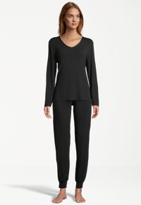 Woman standing barefoot wearing a black long-sleeve V-neck top and matching black jogger-style pants on a white background.