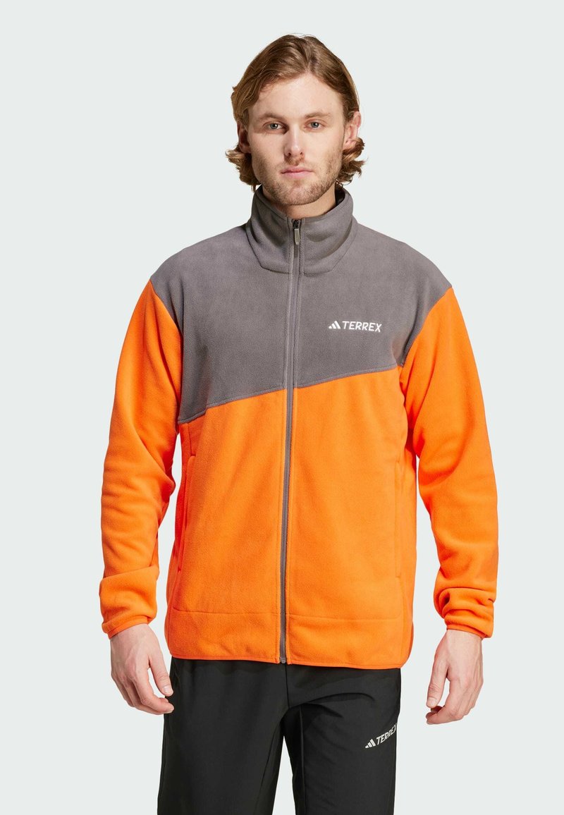 Fleecejakke med lynlås foran; har grå overdel og livlig orange underdel; inkluderer logo detaljer; glat tekstur; fuld lynlås krave.