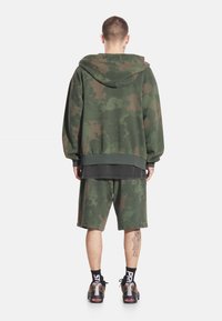 Mężczyzna ubrany w zielono-brązową kamuflażową bluzę z kapturem i szorty, czarne skarpetki z białym napisem oraz wielokolorowe sneakersy, stoi tyłem.