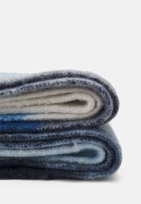 Couverture en laine pliée en couches, présentant des nuances de bleu, gris et crème. Texture douce avec une surface légèrement duveteuse et des bords finis.