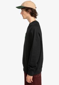 Black sweatshirt with ribbed cuffs and hem, featuring a small green graphic detail. Worn with a tan cap with a green brim.