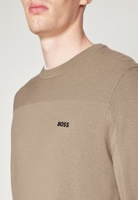 Beige knit sweater featuring a textured body, smooth round neckline, and a small black "BOSS" logo on the chest.