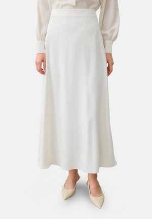 Vrouw draagt een lange, witte, vloeiende rok gecombineerd met beige slingback schoenen met een puntige neus tegen een effen achtergrond.