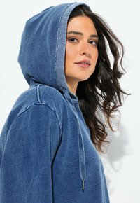 Hoodie sininen denimistämalli, jossa on teksturoitu pinta, nyöri ja korostetut saumat, muotoiltu rentoon istuvuuteen.
