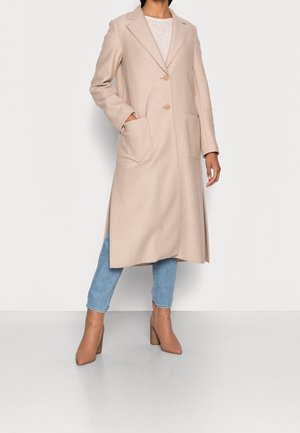 Osoba ubrana w beżowy długi płaszcz z dużymi przednimi kieszeniami, niebieskie dżinsy oraz beżowe botki na obcasie, stojąca na tle białego tła.