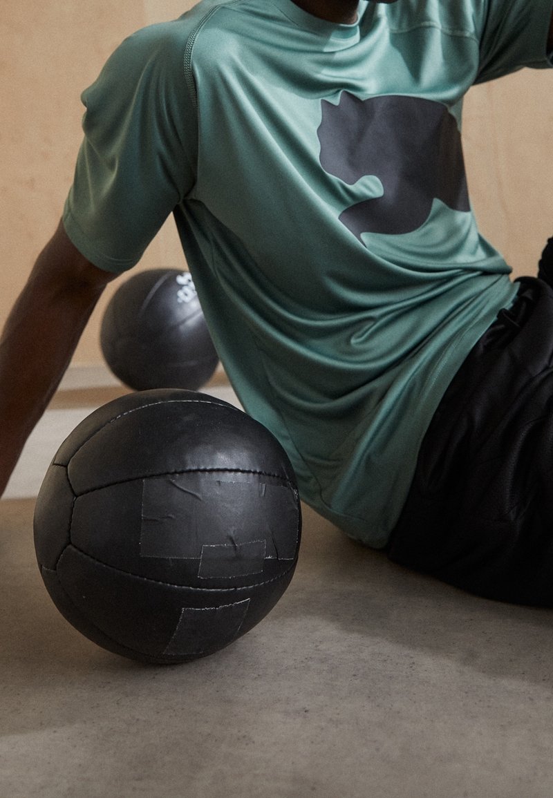 Camiseta deportiva verde con un logotipo gráfico negro, combinada con pantalones cortos negros, junto a una pelota medicinal negra texturizada sobre un suelo de cemento.