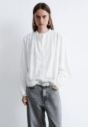 Witte geborduurde blouse met een ronde hals en lange mouwen, gecombineerd met hooggetailleerde grijze jeans en een witte riem.