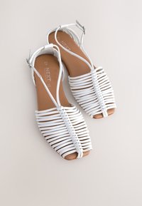 Sandales blanches à brides avec une semelle plate, un design tissé, un bout ouvert et une boucle réglable à la cheville. Texture lisse avec une semelle intérieure en cuir naturel.