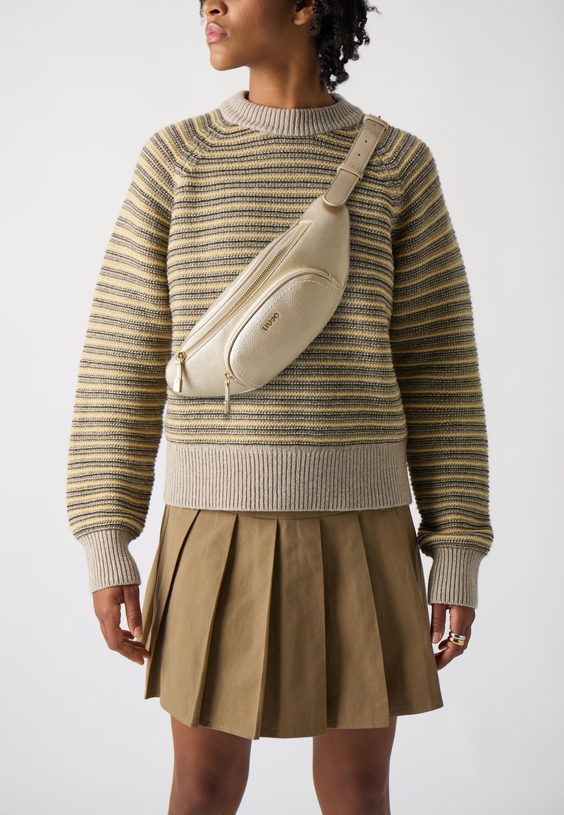 Beige laukku, joka on valmistettu teksturoidusta nahasta, on yllään raidallisen neulepuseron ja laskostetun khaki-hameen päällä, esitellen modernia, rentoa muotoilua.