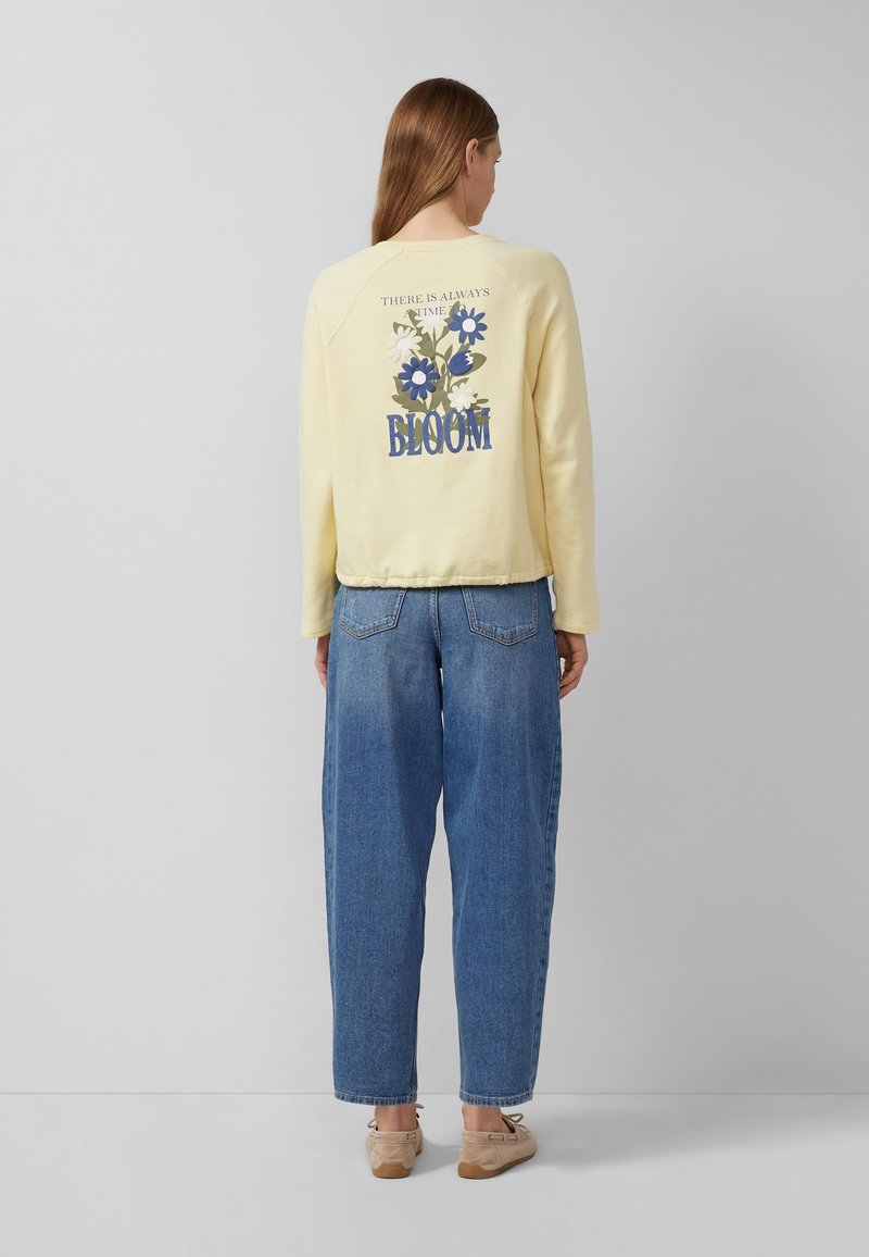 Femme debout de dos portant un pull jaune clair avec des fleurs bleues et blanches et le texte « Il y a toujours du temps pour s'épanouir », ainsi qu'un jean bleu.