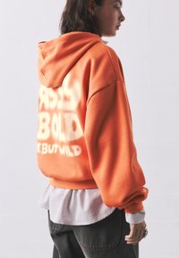Orange oversized hoodie with a front pocket, featuring bold white text on the back. Layered over a light blue striped shirt. Dark jeans visible.