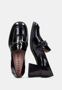 Mocasines de charol negro con tacón cuadrado, que presentan una hebilla metálica decorativa y un acabado suave y brillante.