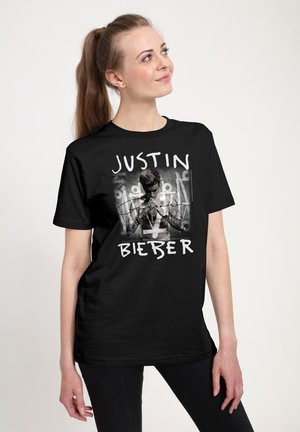 Młoda kobieta w czarnej koszulce z grafiką Justina Biebera, patrząca w górę i lekko się uśmiechająca, stojąca na jasnym, jednolitym tle.