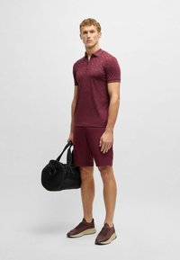Mörkröd pikétröja med ett strukturerat mönster, matchande shorts. Håller en svart gymväska, bär mörkröda och beige sneakers.