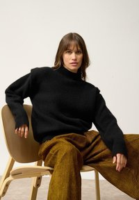 Czarny dzianinowy sweter z golfem i opadającymi ramionami, w połączeniu z brązowymi spodniami w szerokim kroju z pionowymi paskami, siedzący na drewnianym krześle.