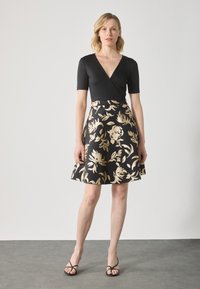 Robe noire drapée avec des manches courtes et un corsage ajusté, présentant une jupe évasée ornée de motifs floraux dorés. Sandales noires fines.