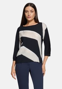 Zwart-crème top met een abstract zigzagpatroon, driekwart mouwen en een ronde halslijn, gemaakt van een glad, lichtgewicht materiaal.