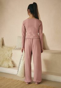 Completo loungewear a coste di un rosa chiaro, con maniche lunghe e pantaloni dalla vestibilità morbida. Il modello tiene un cuscino decorativo in una mano. Dettagli di texture sottili.