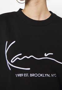 Sudadera negra con el logo bordado en blanco "Kani" y el texto "1989 EST. BROOKLYN, NYC" en la parte delantera, llevada por una persona con un pendiente de aro.