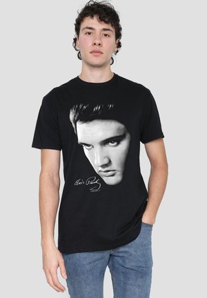 Young man wearing a black t-shirt with a large black-and-white portrait of Elvis Presley and his signature, paired with blue jeans.