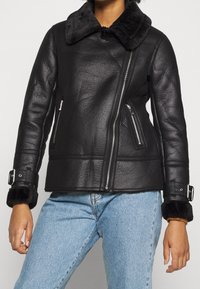 Personne portant une veste en shearling noir avec une fermeture éclair asymétrique, des poignets à boucle et un jean bleu clair, se tenant devant un fond uni.