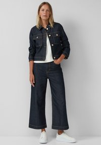 Dunkelblaue Jeansjacke mit goldenen Knöpfen, tailliertem Design und zwei vorderen Taschen. Kombiniert mit weit geschnittenen dunklen Jeans und weißen Sneakern.
