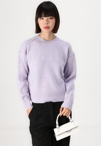 Pull lilas avec un ourlet et des poignets côtelés, présentant une coupe décontractée et une silhouette légèrement oversized. Associé à un petit sac à main blanc.