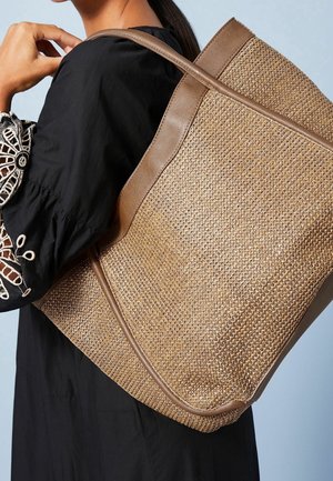 Sac fourre-tout en paille tissée avec des accents en cuir marron, présentant une forme structurée et un haut ouvert, porté à l'épaule.