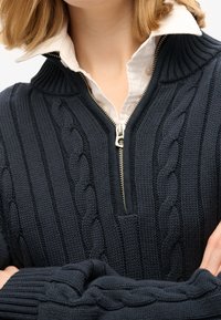 Marineblauer Strickpullover mit hohem Kragen, vorne mit Reißverschluss und strukturierten Zopfmustern. Gerippte Abschlüsse an Ärmeln und Saum.