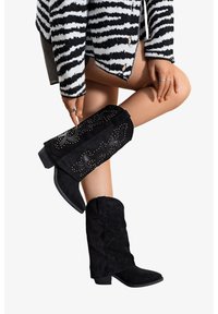 Bottines en daim noir avec des bords courbés, des coutures décoratives et des clous. Talons épais et texture douce.