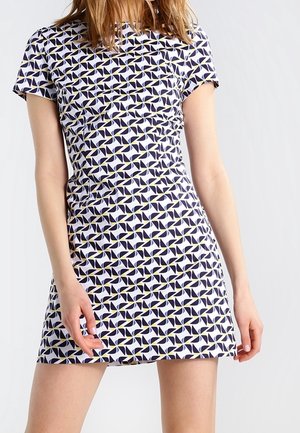 Femme portant une robe à manches courtes, au-dessus du genou, avec un motif géométrique répétitif de moulins blancs et bleu marine avec de petits accents jaunes.