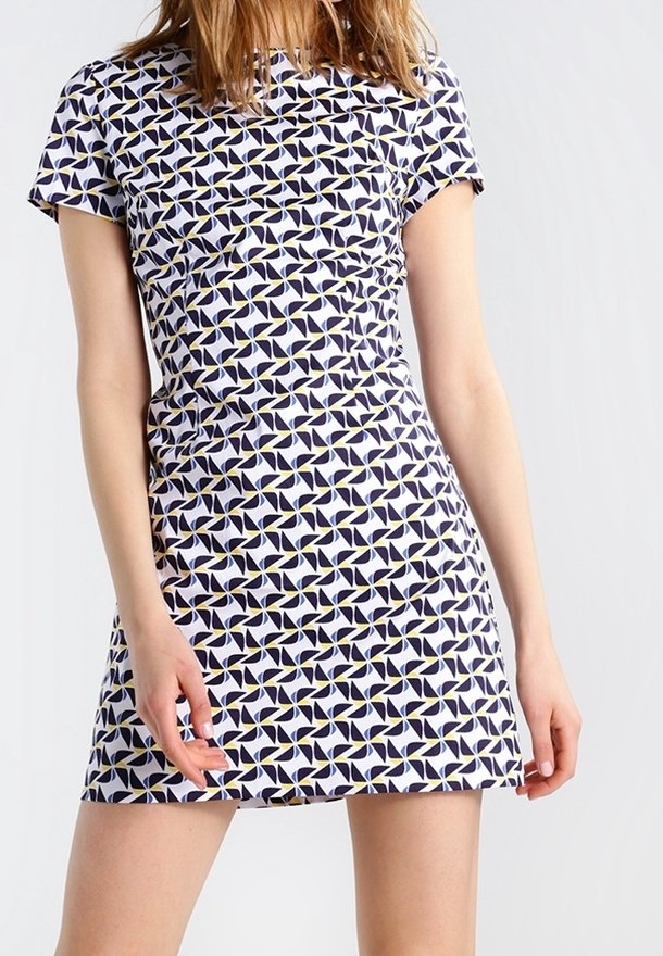 Femme portant une robe à manches courtes, au-dessus du genou, avec un motif géométrique répétitif de moulins blancs et bleu marine avec de petits accents jaunes.