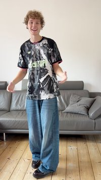 Svartvit mönstrad skjorta med en grön logotyp, kombinerad med löst sittande ljusblå jeans och svarta skor, i ett enkelt inomhusutrymme.