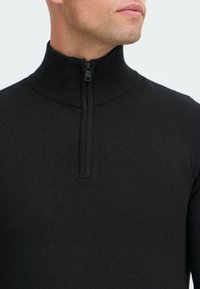 Sweater noir à zip avec un col haut et une texture côtelée. Dispose d'une fermeture éclair d'un quart de longueur et d'un design ajusté.