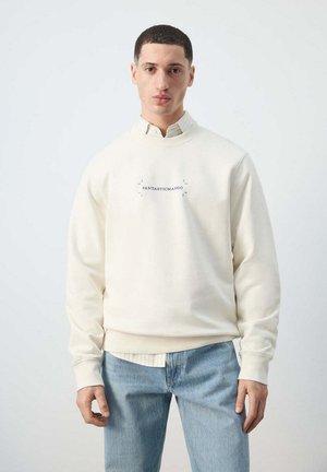 Joven con sudadera color crema con texto "FANTASTICMANGO", camisa de rayas claras debajo y vaqueros azules, de pie frente a un fondo liso.