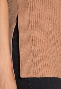 Primo piano di un maglione a coste di colore marrone chiaro con spacco laterale, indossato sopra pantaloni neri, che mostra parte del braccio e della vita dell'indossatore.