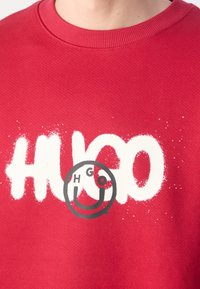 Chemise rouge avec texte blanc style peinture en spray "HUGO" et un visage souriant noir avec des yeux rouges et noirs superposés sur le texte.