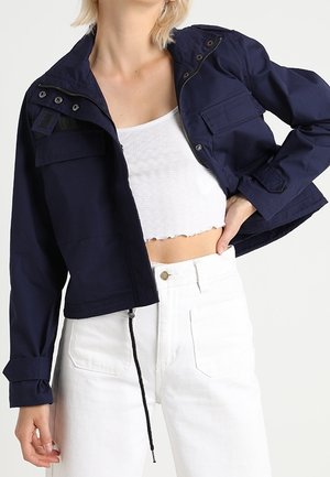 Vrouw draagt een marineblauw cropped jack over een wit geribd cropped topje en een witte broek met hoge taille tegen een effen achtergrond.