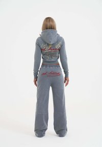 Vrouw in een blauw-grijze hoodie en wijdere broekset met rode en gele borduurschrift gezien vanaf de achterkant op een witte achtergrond.
