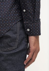 Czarna koszula z długim rękawem w małe niebieskie, białe i rdzawoczerwone kropki. Posiada zapięcie na guziki przy mankietach i zestawiona jest z ciemnoniebieskimi dżinsami.